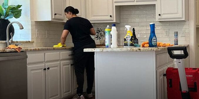 Professional cleaner wiping down a kitchen counter in a modern home, with cleaning supplies and equipment placed on the island.
